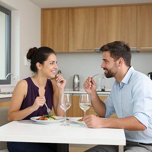 Photograph of a smiling couple dining in a modern kitchen, both holding forks, with wooden cabinets and wine glasses on the table.