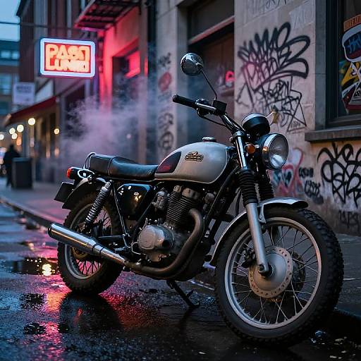 Photograph of a classic silver motorcycle with chrome exhaust pipes, parked on a wet urban street at night, under red neon signs and graffiti-covered buildings.