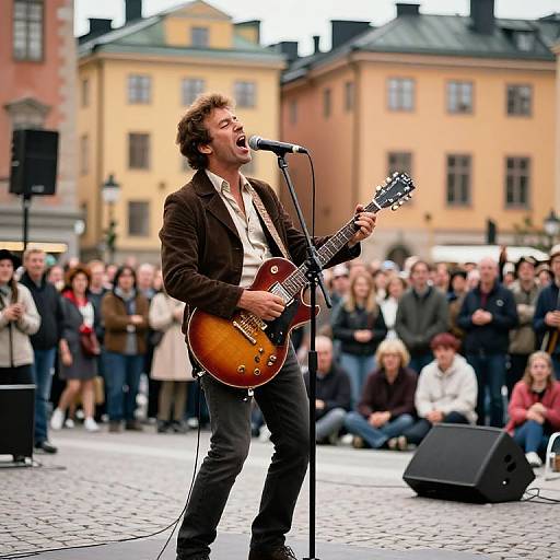 Photograph of a passionate male singer-guitarist with brown hair and beard, wearing a brown blazer, singing into a microphone on a cobble