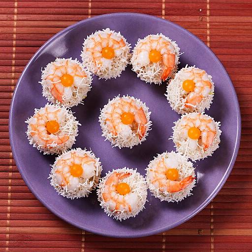 Photograph of eight sushi balls with shrimp and egg on a dark blue plate, on a red bamboo mat.