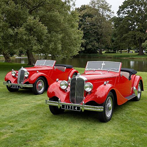 Photograph of two bright red vintage convertible cars with chrome grilles and black roofs, parked on lush green grass near a serene lake with large trees in