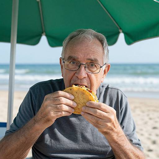 Photograph of an elderly man with gray hair and glasses, wearing a dark shirt, eating a sandwich under a green beach umbrella, with the ocean and