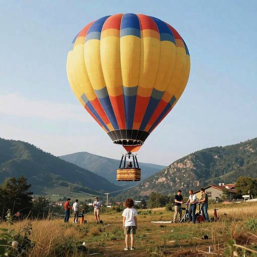 Photograph of a large, striped yellow, red, and blue hot air balloon ascending over a grassy field with mountains and people in the background on