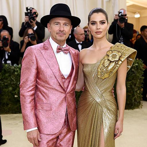 Photograph of a man in a pink suit and black hat, and a woman in a gold one-shoulder gown, posing on a red carpet