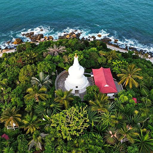 Aerial photograph of a white temple with a red roof, surrounded by lush green palm trees, overlooking a rocky ocean shoreline.