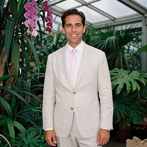 Photograph of a smiling man in a white suit with a pink tie, standing in a lush greenhouse filled with purple orchids and green plants.