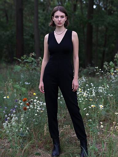Photograph of a young woman with fair skin, brown hair, wearing a sleeveless black jumpsuit and black boots, standing in a lush, floral