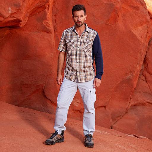 Photograph of a bearded man with short dark hair, wearing a plaid shirt, white pants, and black shoes, standing against a vivid red