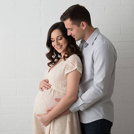 Photograph of a pregnant woman in a lace dress, smiling, with a man in a light blue shirt, gently embracing her from behind, standing against