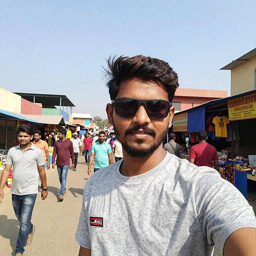 Photograph of a bearded Indian man with dark hair, wearing sunglasses, gray t-shirt, and jeans, standing in a busy outdoor market under a