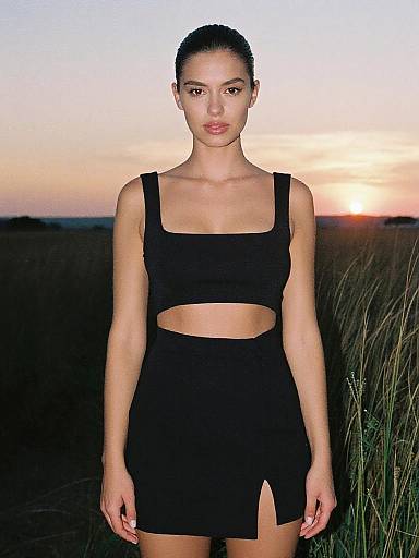 Photograph of a young woman with dark hair in a bun, wearing a black crop top and skirt with a front slit, standing in a grassy