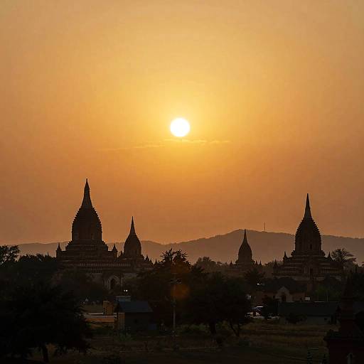 Sunset silhouette of ancient temples with pointed spires against a golden-orange sky, hills in the background, photograph.