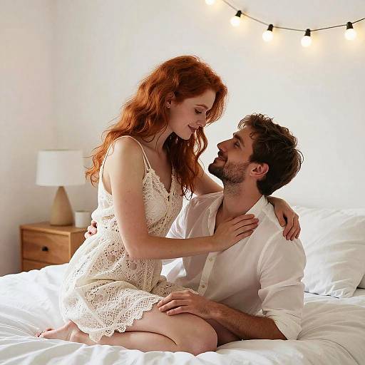 Photograph of a red-haired woman in a white lace dress and a bearded man in a white shirt, sitting on a bed, gazing at