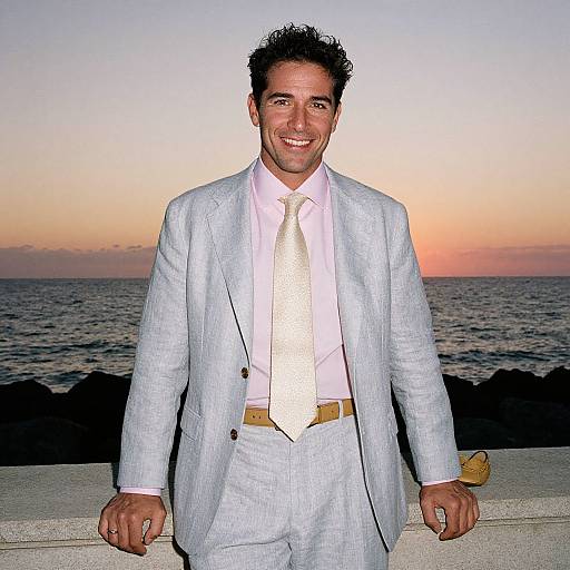 Photograph of a smiling man with short curly hair, wearing a light gray suit, white shirt, and gold tie, standing against a sunset ocean backdrop