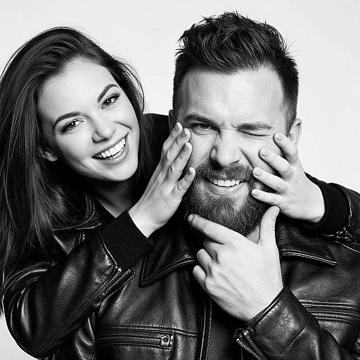 Black-and-white photograph of a smiling woman with long dark hair, playfully touching the face of a bearded man in a leather jacket. White background