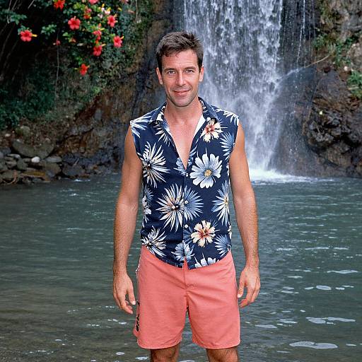 Photograph of a smiling, short-haired man in a black floral shirt and orange shorts, standing in front of a waterfall with lush greenery and red