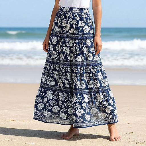 Photograph of a person's lower body in a white top and a long, blue floral skirt standing barefoot on a sandy beach with the ocean and