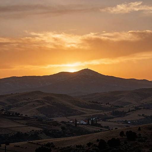 Photograph of a picturesque sunset over rolling hills, with a silhouetted mountain peak and warm, orange-tinged sky, and small village