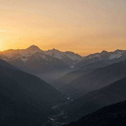 Photograph of a mountain range at sunset, with snow-capped peaks glowing under a golden-orange sky, dark forested valleys in the foreground, and