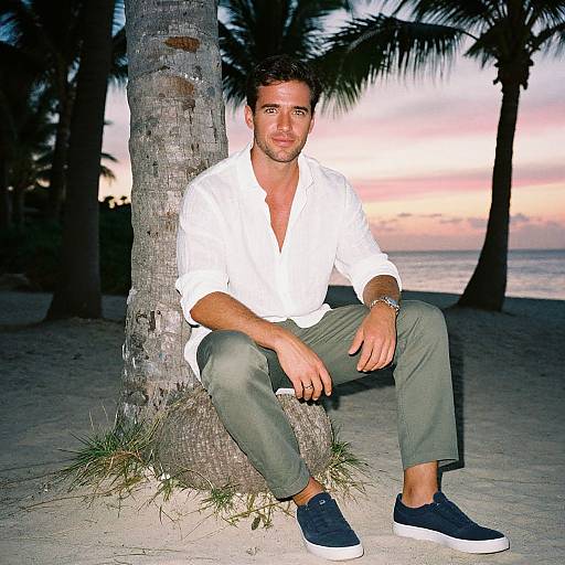 Photograph: Handsome man with short dark hair, white shirt, green pants, and blue sneakers, sitting by a palm tree on a beach at