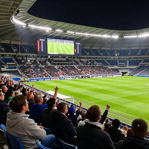 Photograph of a packed stadium with cheering fans, bright green field, and large screen showing a soccer match. Players in action, fans raising arms in