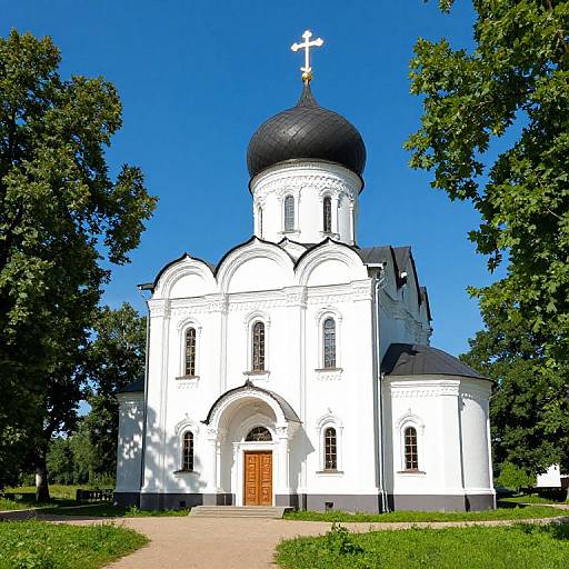 Photograph of a white Orthodox church with a black dome and cross, set amidst lush green trees under a clear blue sky.