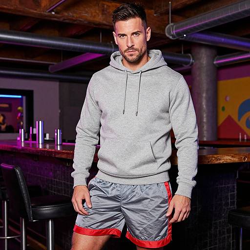 Photograph of a muscular, bearded man with short dark hair, wearing a gray hoodie and gray shorts with red trim, standing confidently in a modern