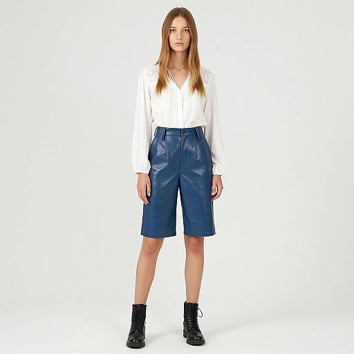 Photograph of a young woman with long brown hair, wearing a white blouse, blue leather shorts, and black ankle boots, standing against a plain white