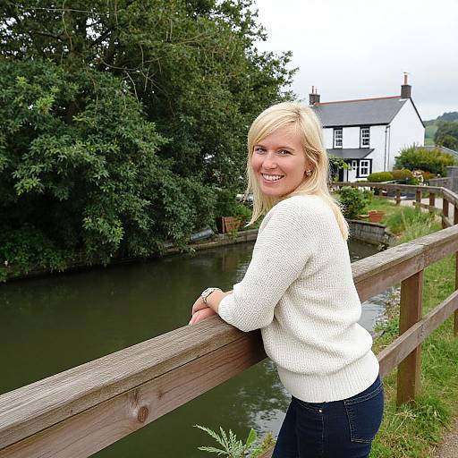 Blonde woman in white sweater and blue jeans smiles while leaning on wooden fence by serene river and white cottage.