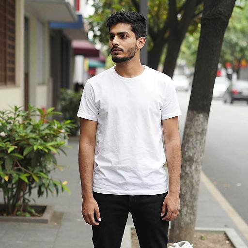 Photograph of a young, handsome man with short black hair and beard, wearing a white t-shirt and black pants, standing on a sunlit urban