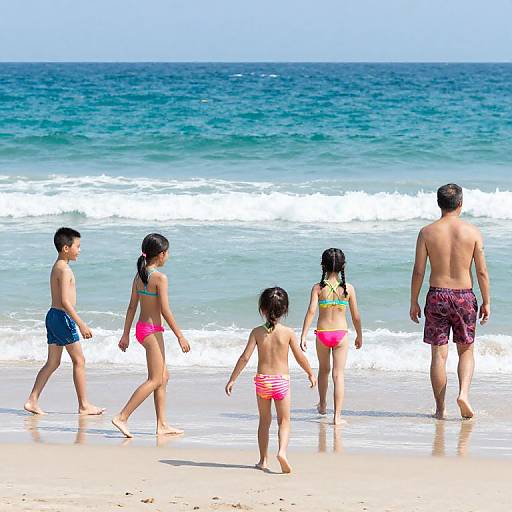 Photograph of a family with four children and one adult walking on a sunny beach with blue ocean waves in the background.