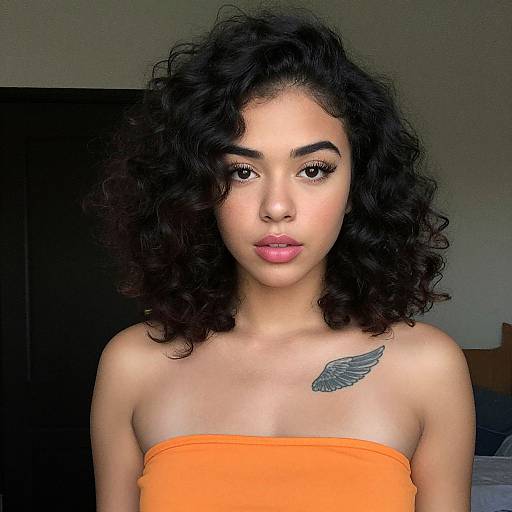 Photograph of a young woman with curly black hair, light brown skin, and a small wing tattoo on her right shoulder, wearing an orange strapless