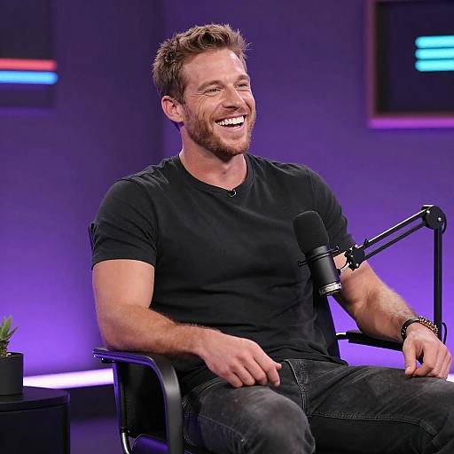 Photograph of a smiling, bearded white man with short brown hair, wearing a black t-shirt and jeans, seated on a chair with a microphone