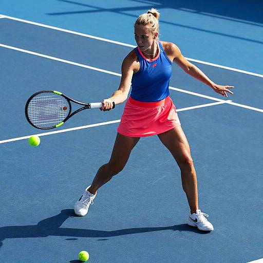 Photograph of a blonde woman in a blue tank top and pink skirt, playing tennis on a blue court, hitting a yellow ball with a black racket