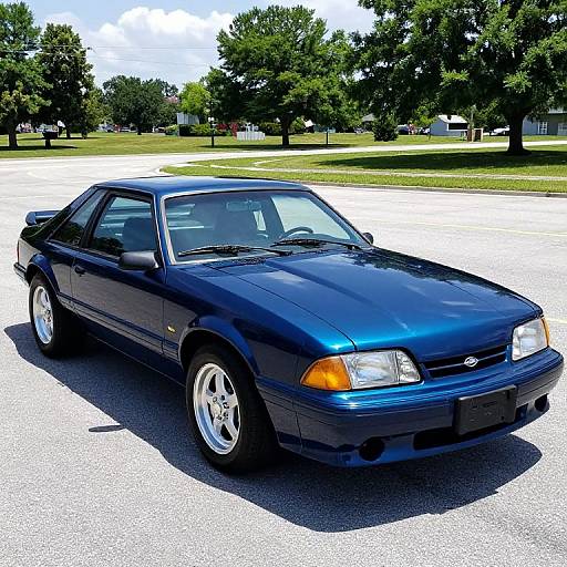 Photograph of a shiny, blue, 90s Chevrolet Camaro parked on a sunlit, empty lot with green trees and a clear sky in