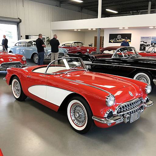 Photograph of a vintage car showroom featuring a bright red convertible with white accents, chrome grille, and white-walled tires, surrounded by other classic cars