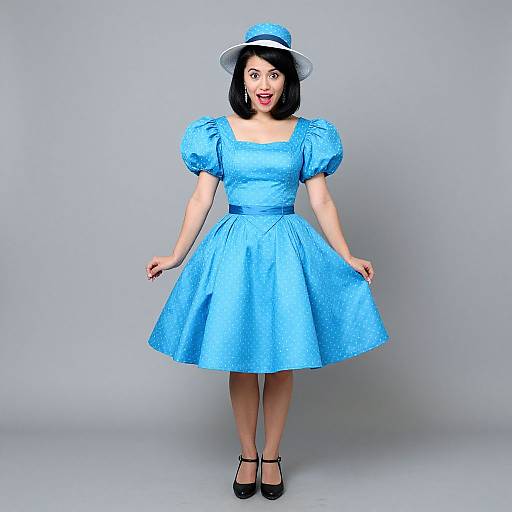 Photograph of a smiling Asian woman in a bright blue, puffy-sleeved, knee-length dress and matching hat, standing against a plain gray