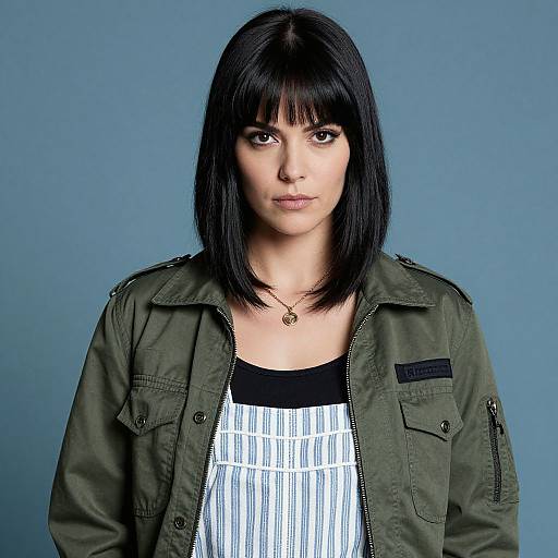 Photograph of a woman with straight black bob haircut, green military jacket, white striped top, and pendant necklace, against a blue background.