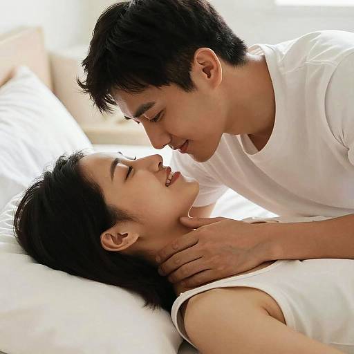 Photograph of an Asian couple in white shirts, lying in bed, gazing into each other's eyes, smiling intimately, with soft, natural light