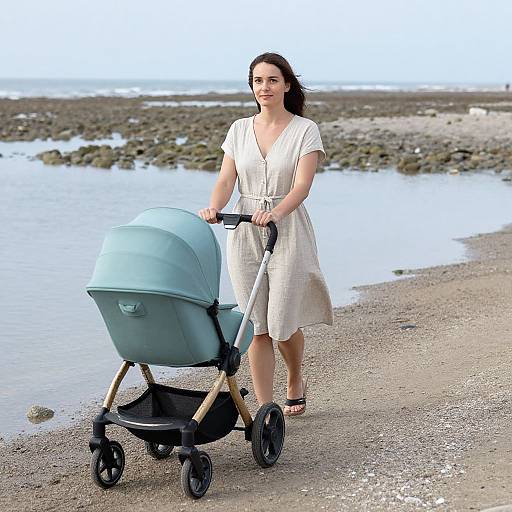 Photograph of a smiling woman with brown hair, wearing a white dress, pushing a light blue stroller on a rocky beach.