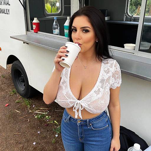 Photograph of a dark-haired woman with large breasts, wearing a white lace crop top and blue jeans, drinking from a white cup at a food truck