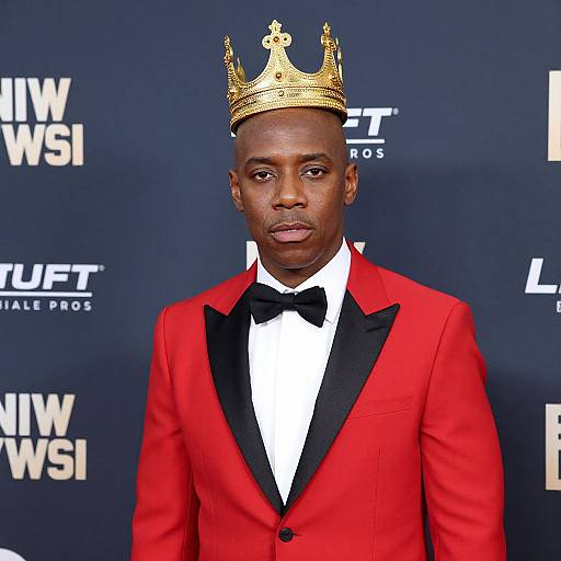 Photograph of a Black man in a red tuxedo with black lapels, black bow tie, and a gold crown, standing against a blue