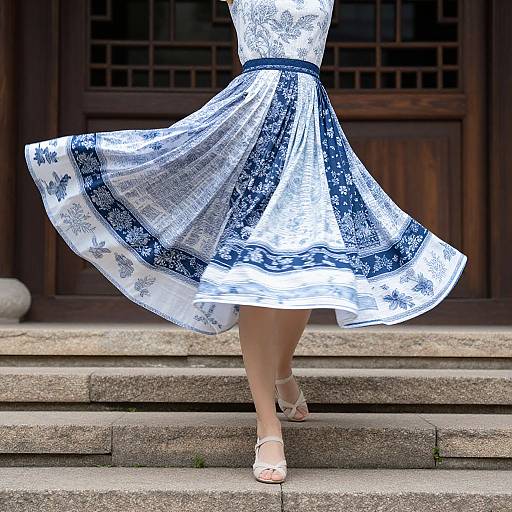 Photograph of a woman's lower body in a white and blue floral dress, mid-step on stone stairs, wearing white sandals.
