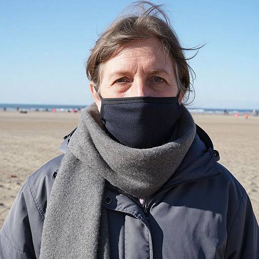 Photograph of middle-aged man with brown hair, wearing black mask and gray scarf, in black coat, standing on sunny beach.