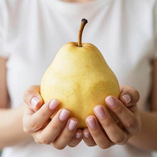 Photograph of a person's hands with pink nail polish, holding a yellow pear with a brown stem, against a bright white background.