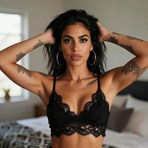 Photograph of a tattooed, dark-haired woman with hoop earrings, wearing a black lace bralette, standing with hands in hair, in a softly
