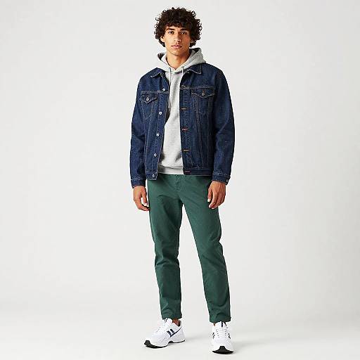 Photograph of a young man with curly hair, wearing a blue denim jacket, white hoodie, green pants, and white sneakers, standing against a white