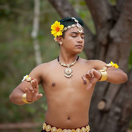 Photograph of a shirtless, dark-skinned man with yellow flower headpiece, gold jewelry, and armlets, dancing outdoors against a forest background
