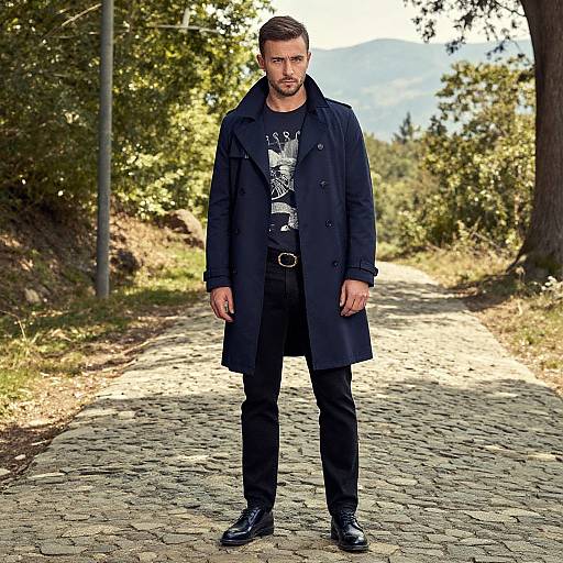 Photograph of a serious, bearded man with short brown hair, wearing a dark navy coat, black graphic shirt, black pants, and boots,