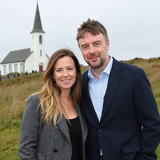 Photograph of a smiling couple standing outdoors in front of a white church with a black steeple, wearing formal attire.